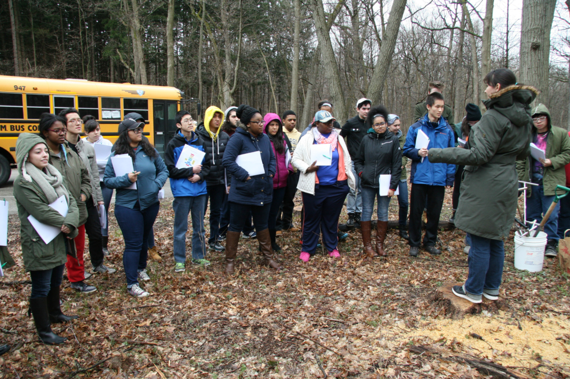 Group of students outside school bus surrounding teacher in woods